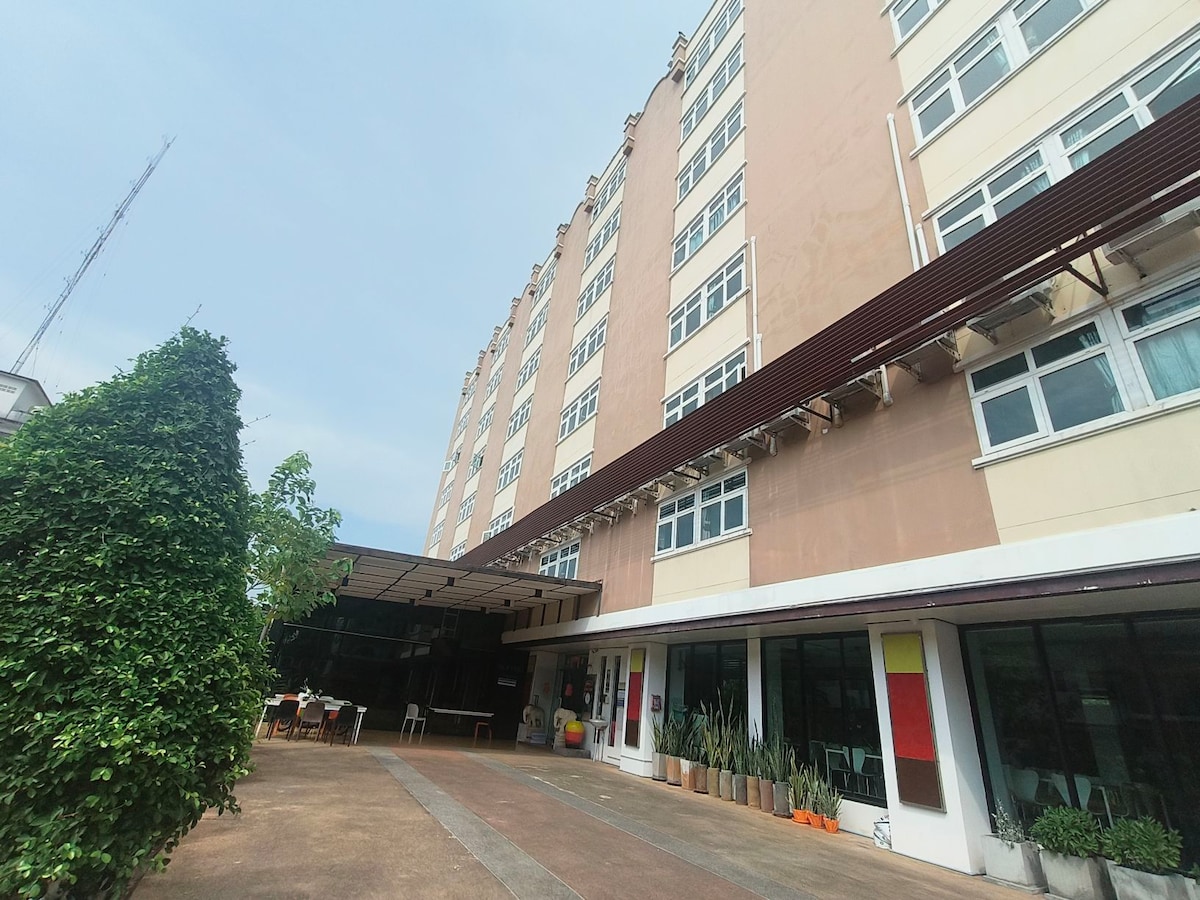 The exterior of a multi-story building is visible, featuring large windows and a covered outdoor seating area. Lush greenery is present in the foreground, with potted plants arranged along the ground. The structure's neutral facade contrasts with colorful elements incorporated into the design.