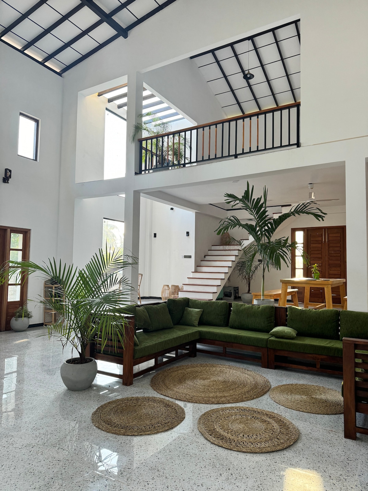 A spacious living area features a large green sofa arranged around natural fiber rugs. Light filters through large windows and a clear-glass ceiling, illuminating indoor plants throughout. A staircase leads to the second floor, while wooden accents and a dining area create a cohesive space.