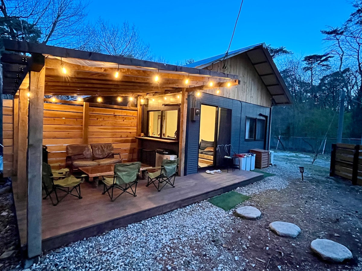 An outdoor seating area features cozy green chairs arranged on a wooden deck, illuminated by string lights overhead. A bar counter is visible, and an entrance leads into the interior space, surrounded by a tranquil natural setting.
