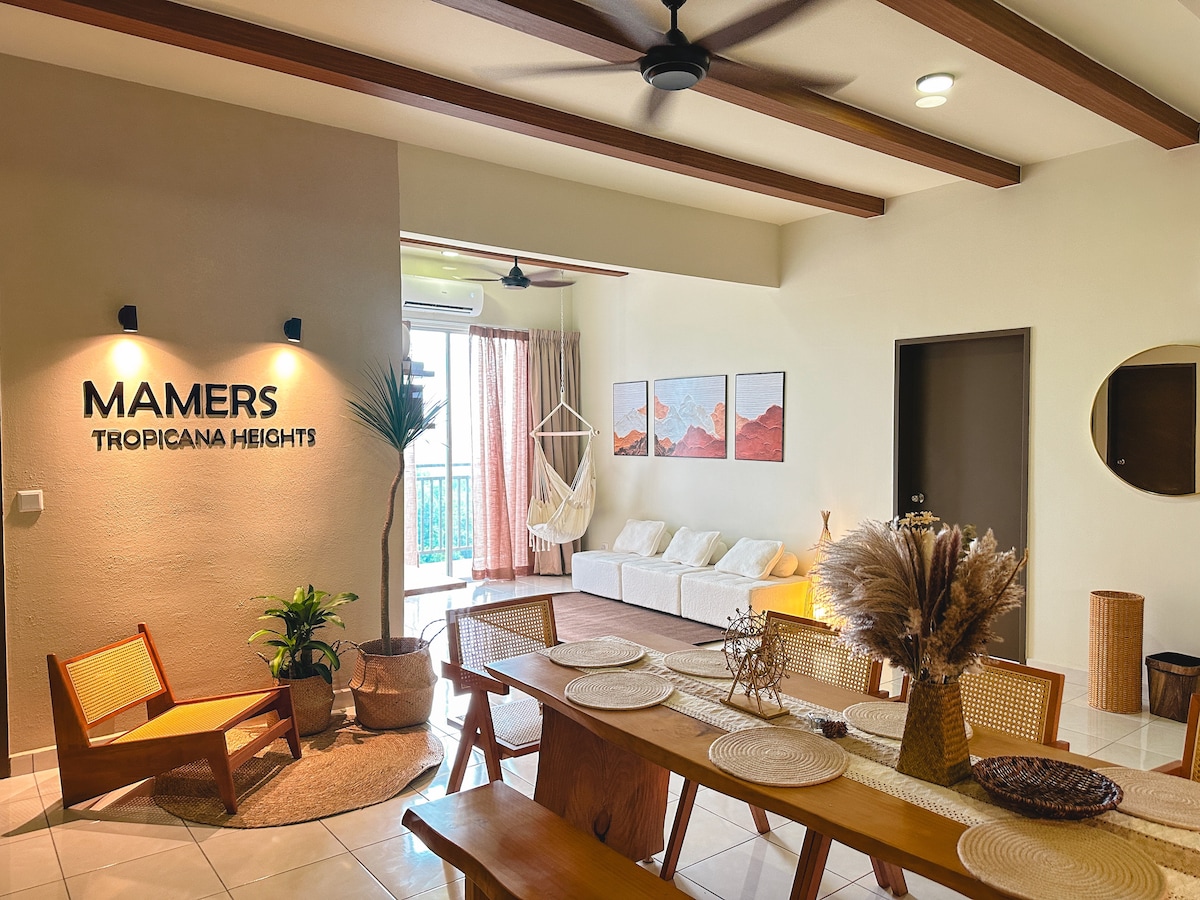 A bright living space features wooden beams and contemporary decor. Natural light enters through large windows, illuminating a cozy seating area with a neutral palette. A long dining table is adorned with woven placemats and a decorative arrangement of dried plants.