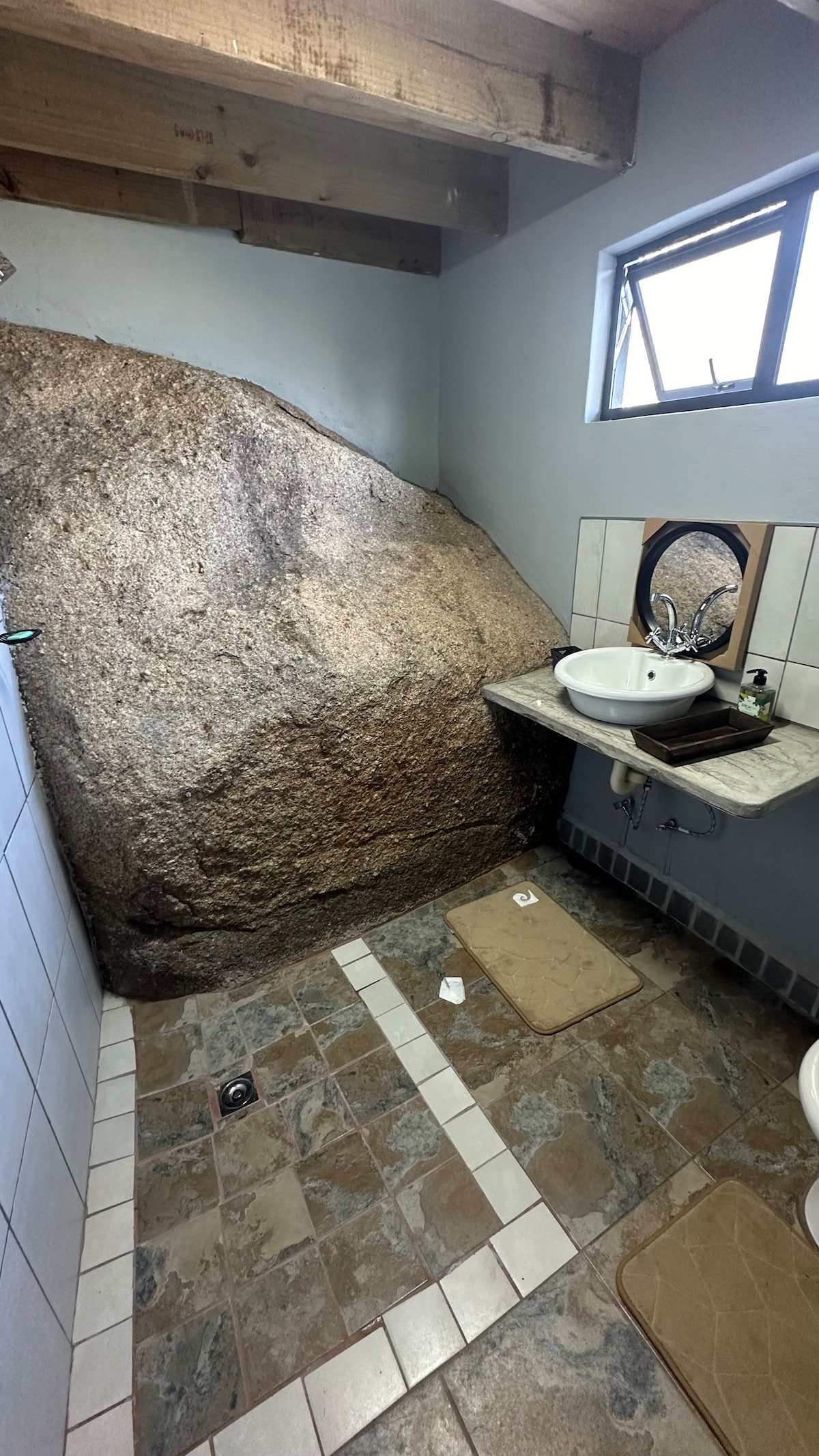 A unique bathroom features a large natural rock as an integral part of the design, contrasting with smooth tiles. A sink with a round mirror is positioned on a countertop against the rock, while neutral-toned mats provide comfort on the floor.