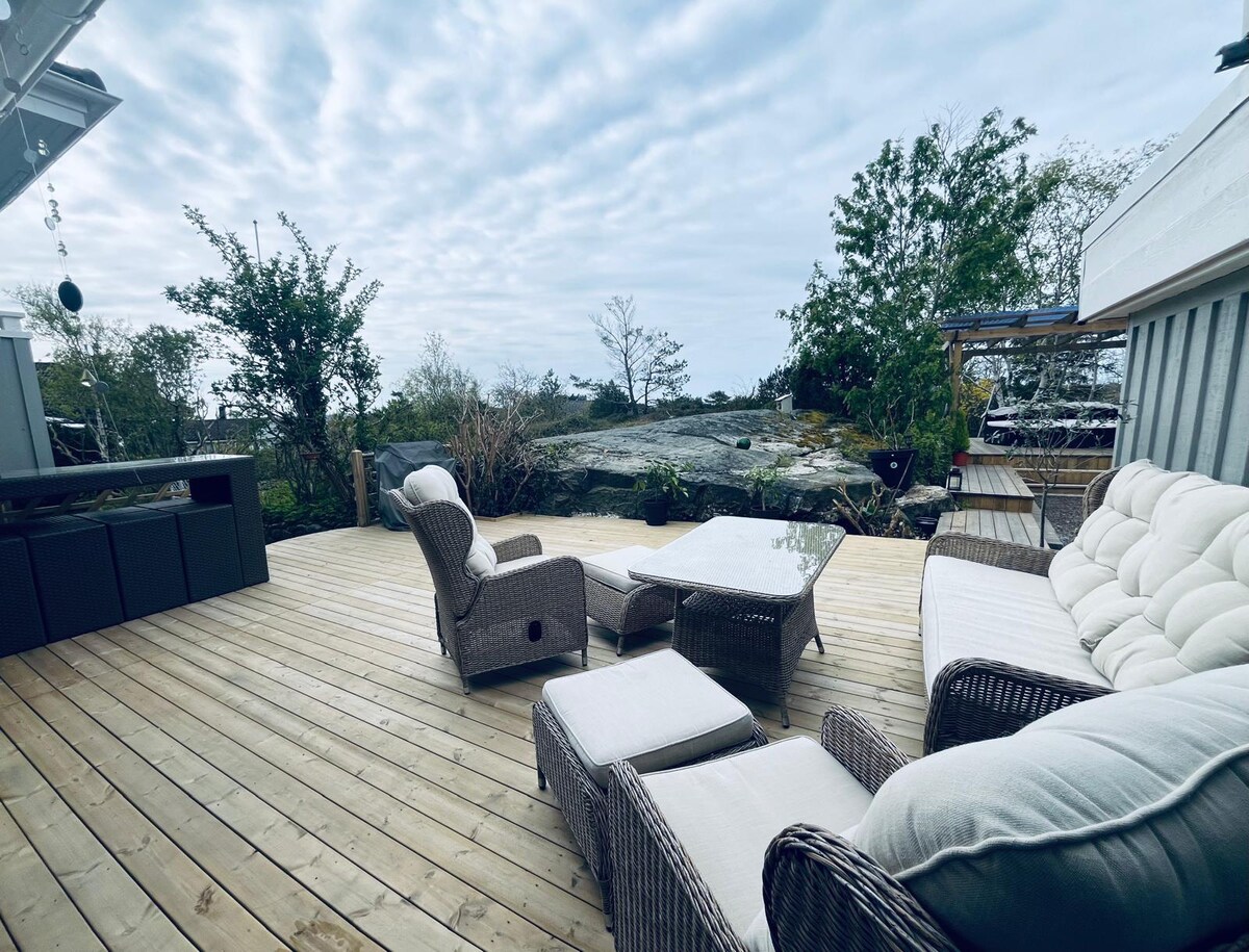 An outdoor terrace features wooden decking, complemented by a comfortable seating area composed of woven furniture. A coffee table sits at the center, surrounded by soft cushions. Greenery and natural rock formations are visible in the background, enhancing the tranquil setting.