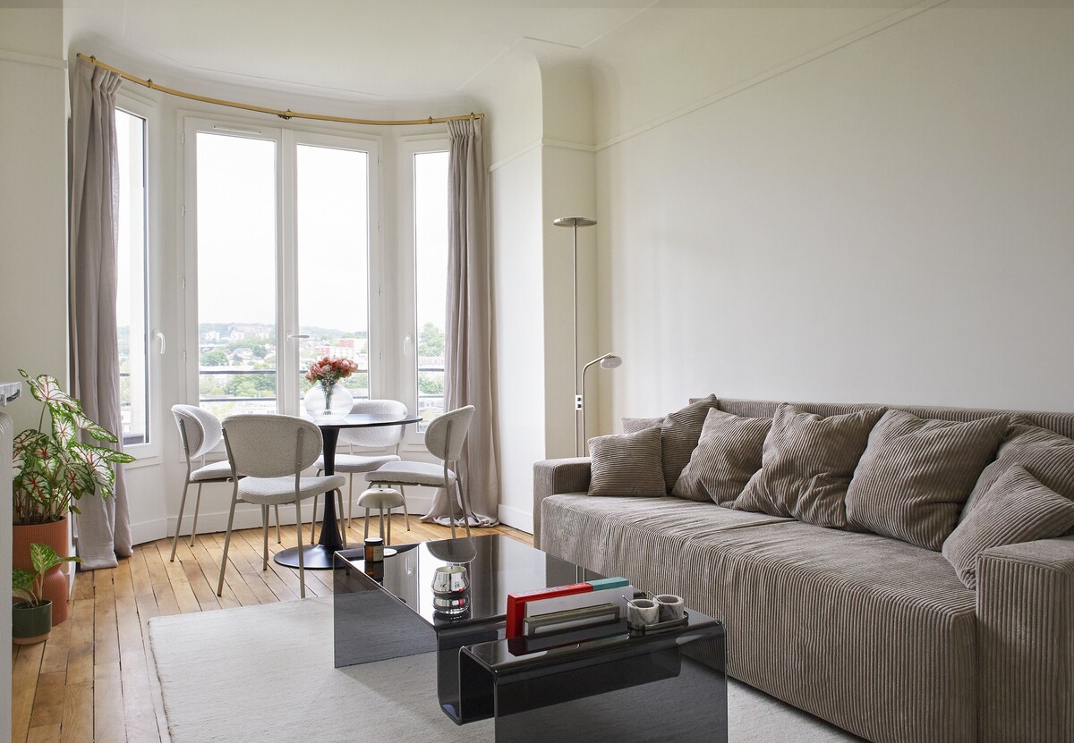 A comfortable living area is highlighted by a spacious sofa and a modern coffee table. A round dining table with four chairs sits near large bay windows, offering natural light. A potted plant adds a touch of greenery to the room, enhancing the overall ambiance.