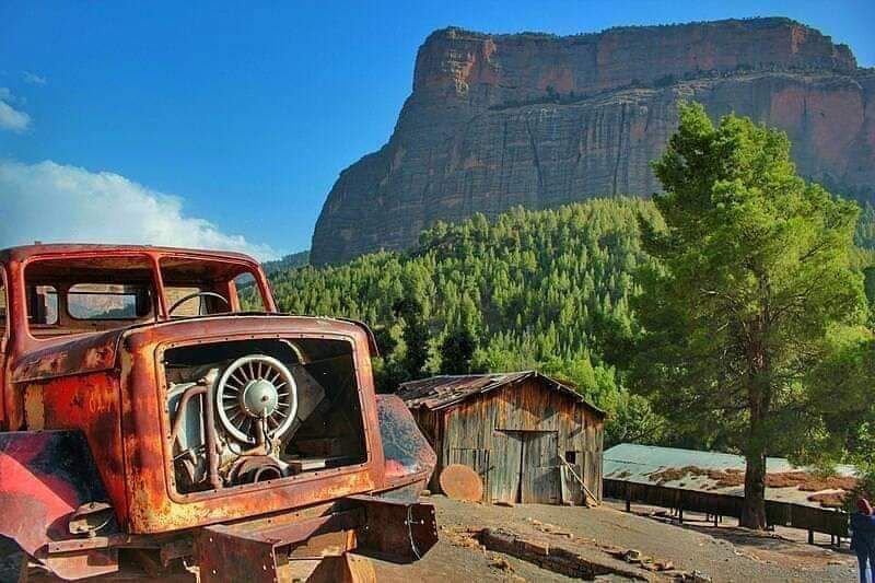 An old, rusty truck is positioned in the foreground, with the engine visible and framed against a backdrop of a large mountain. A weathered wooden structure stands nearby, surrounded by lush greenery and tall trees, under a clear blue sky with scattered clouds.