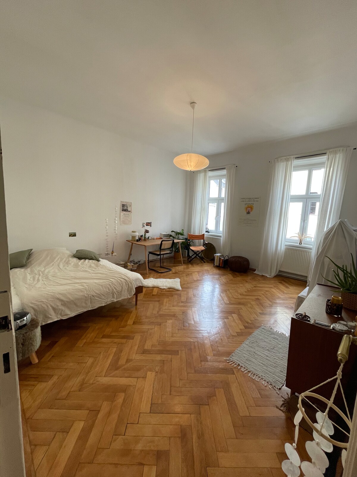 A spacious room features herringbone wooden flooring and a large window dressed with light curtains. A light fixture hangs from the ceiling. A bed is positioned against one wall, while a small desk and a plant are situated nearby. Decorative elements enhance the minimalist decor.