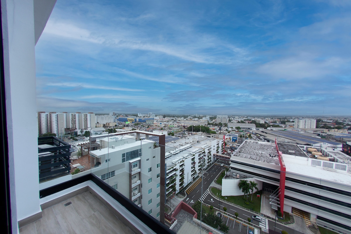 The balcony offers a panoramic view of the surrounding cityscape, featuring multiple buildings and roads stretching towards the horizon. The sky is filled with soft clouds, creating a serene backdrop. A glass railing enhances the open feeling of the space.