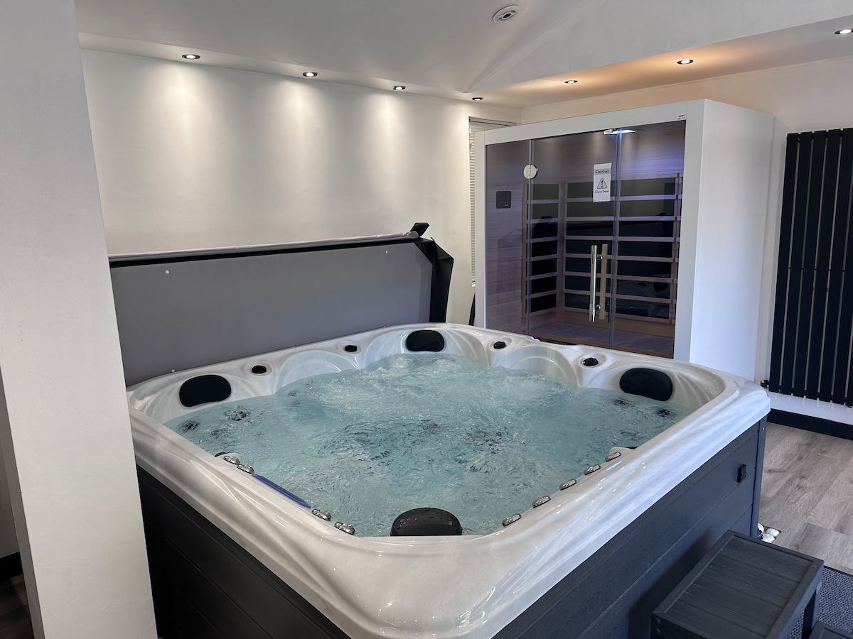 An indoor hot tub is positioned prominently within a well-lit space. Water bubbles gently, reflecting the soft illumination from recessed lighting above. Adjacent, a sauna is visible with a glass door, completing the tranquil atmosphere designed for relaxation.