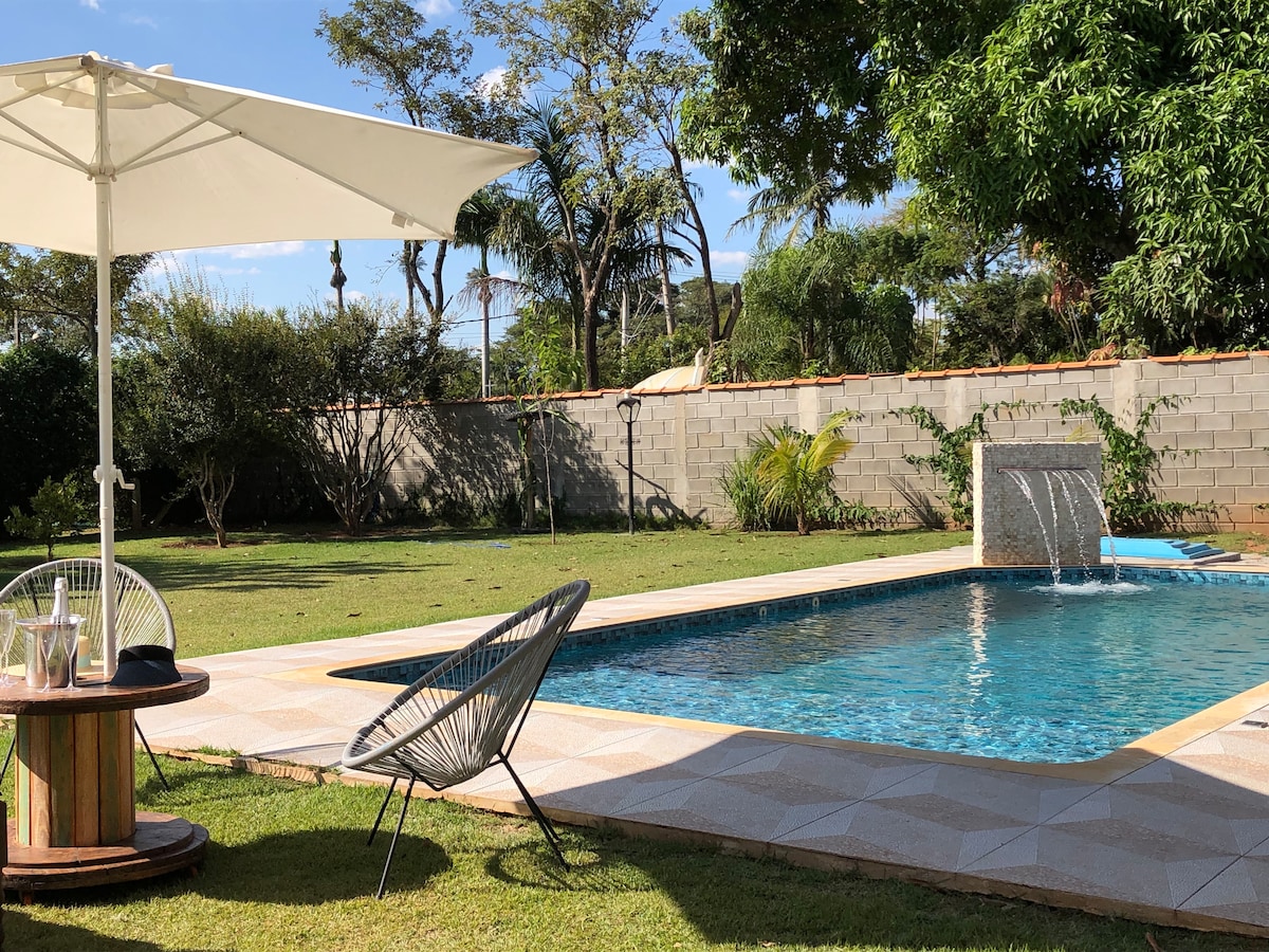 A serene outdoor area features a shimmering pool with a cascading waterfall. A shaded umbrella stands over a comfortable chair beside the pool, and lush greenery provides a natural backdrop. The well-maintained lawn offers ample space for relaxation and outdoor activities.