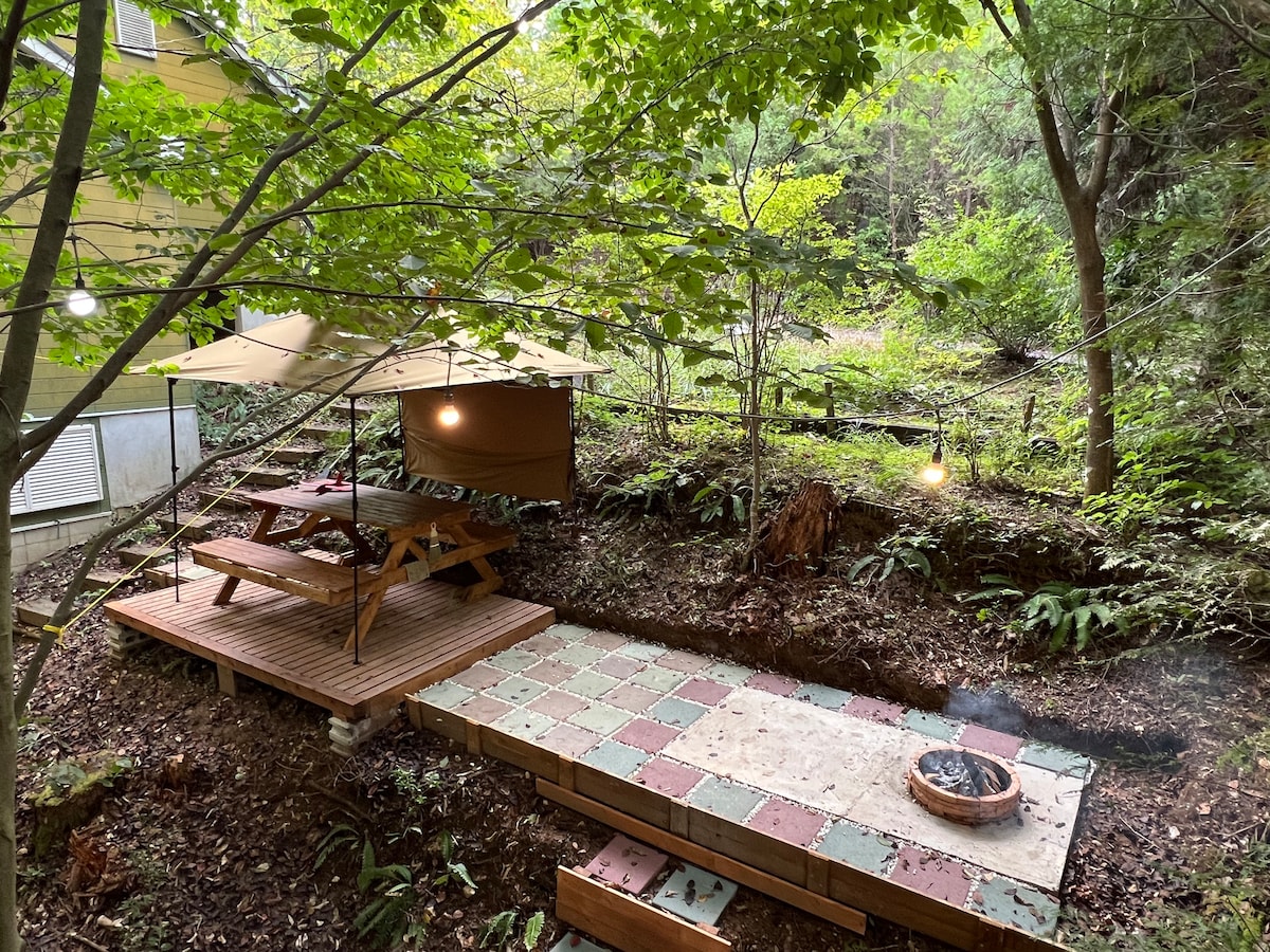 A cozy outdoor BBQ area is set among the trees, featuring a wooden picnic table and benches. Soft lighting is provided by hanging lamps, while a fire pit sits on a square stone seating area, blending harmoniously with the surrounding nature.