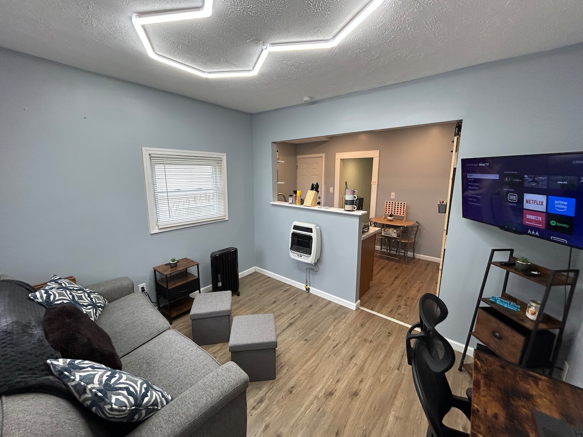 The open living area features a comfortable gray sofa, two small ottomans, and a wall-mounted television displaying various streaming options. A dining area is visible through an archway, complemented by a small desk and chair. A bright, modern ceiling light enhances the inviting space.