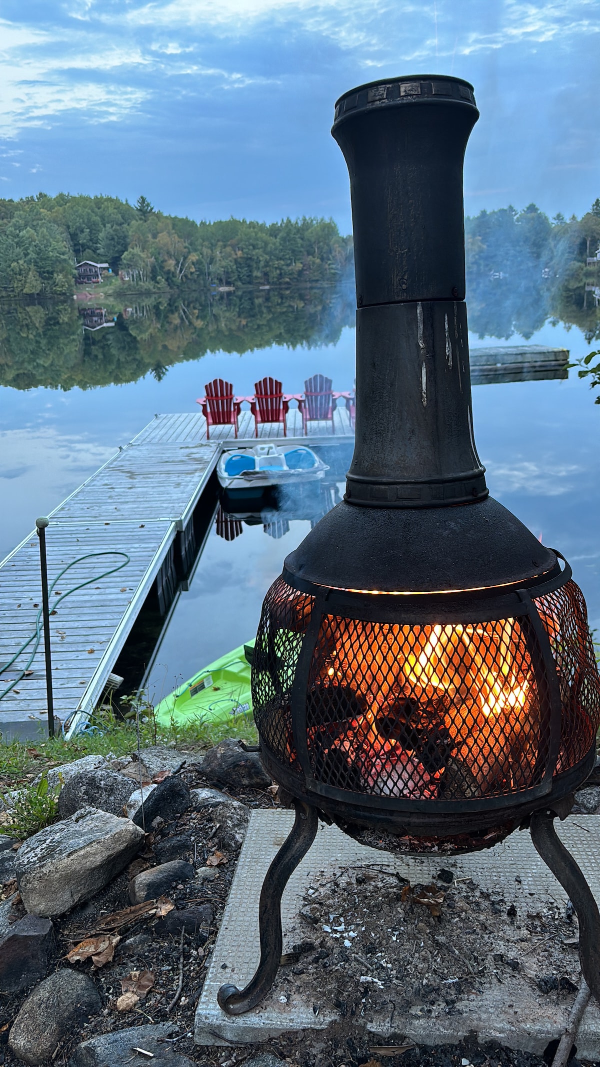 A fire pit stands prominently in the foreground, emitting a warm glow, while a wooden dock leads to the serene lake in the background. Red Adirondack chairs are lined up along the dock, adjacent to pedal boats and kayaks, all surrounded by lush greenery.