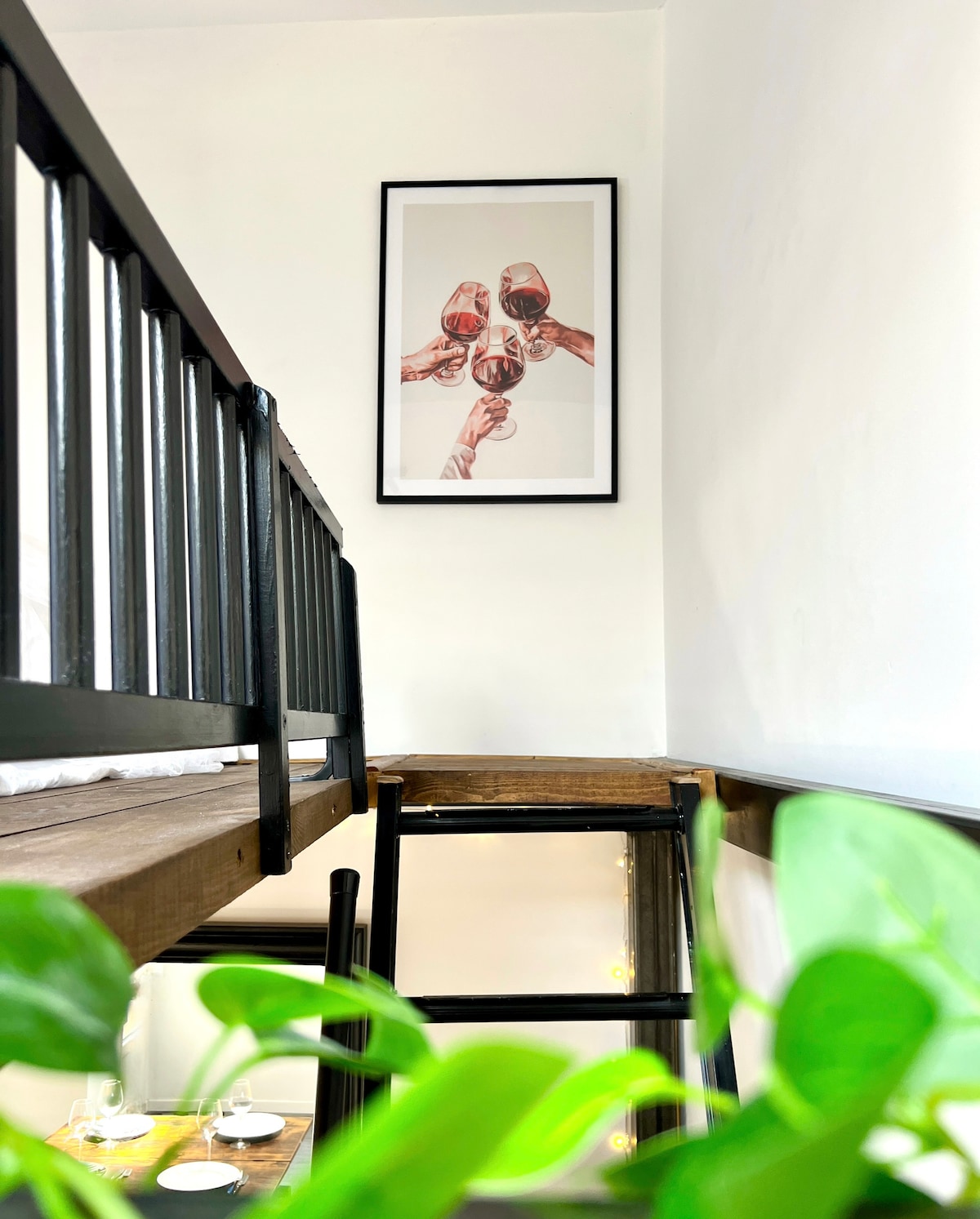 A mezzanine is seen from below, featuring a wooden sleeping area. A framed artwork depicting hands holding wine glasses hangs on the wall above. Green plants are visible in the foreground, adding a touch of freshness to the scene.