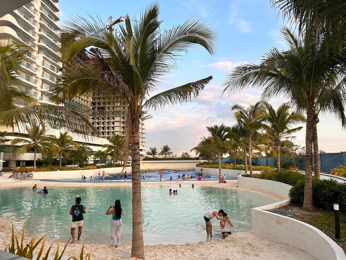 A man-made beach area features soft white sand and azure waters, surrounded by palm trees swaying gently. Guests of various ages can be seen enjoying the water, with some engaging in playful activities along the shore. The scene is complemented by a backdrop of modern buildings.