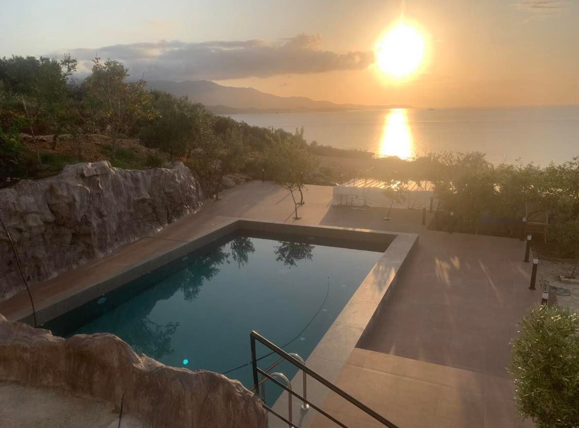 The swimming pool is framed by natural stone features and overlooks a serene sea view at sunset. The water reflects the golden hues of the sky, while lush greenery surrounds the area, creating a peaceful outdoor setting.