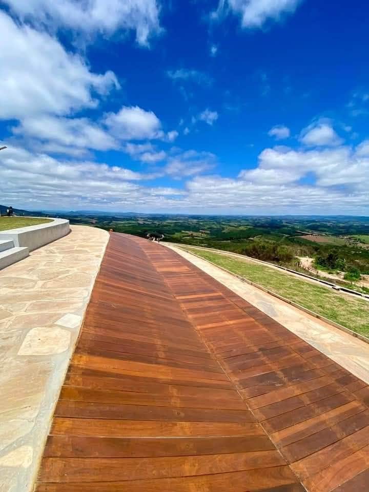 A wooden pathway curves towards the horizon, offering expansive views of green hills and a bright blue sky. Soft white clouds drift above, enhancing the serene landscape. The pathway is bordered by a paved area, inviting guests to explore the scenic surroundings.