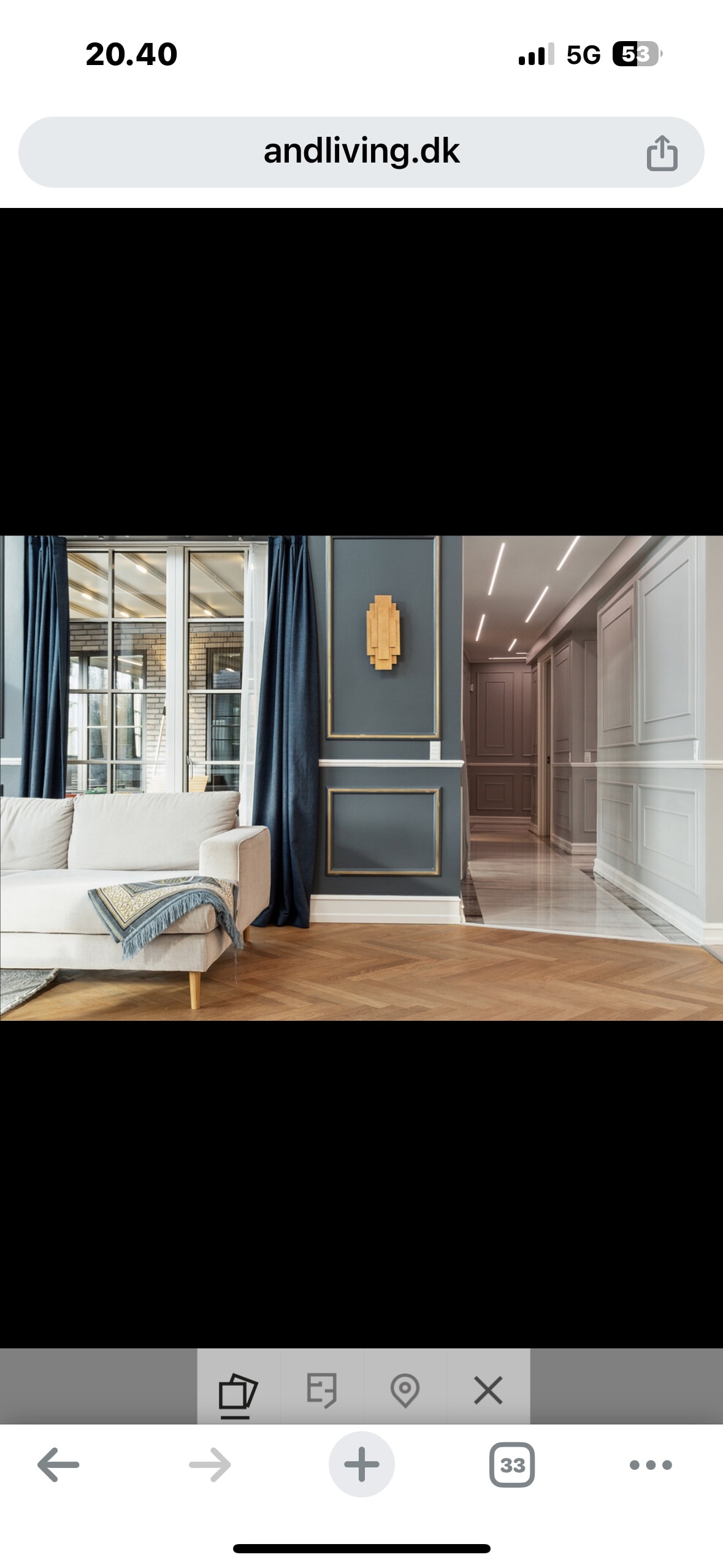 An elegant living area is showcased with a light-colored sofa draped with a soft throw. Dark curtains frame large windows, allowing natural light to illuminate the space. A spacious hallway with polished flooring and recessed lighting extends in the background, accented by decorative wall paneling.