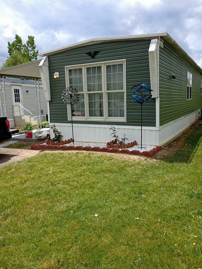 The exterior of a green mobile home is visible, featuring large windows with a grid pattern. Decorative garden elements, including two wind spinners and a landscaped flower bed, are positioned in front. A well-maintained lawn surrounds the structure, contributing to a neat appearance.