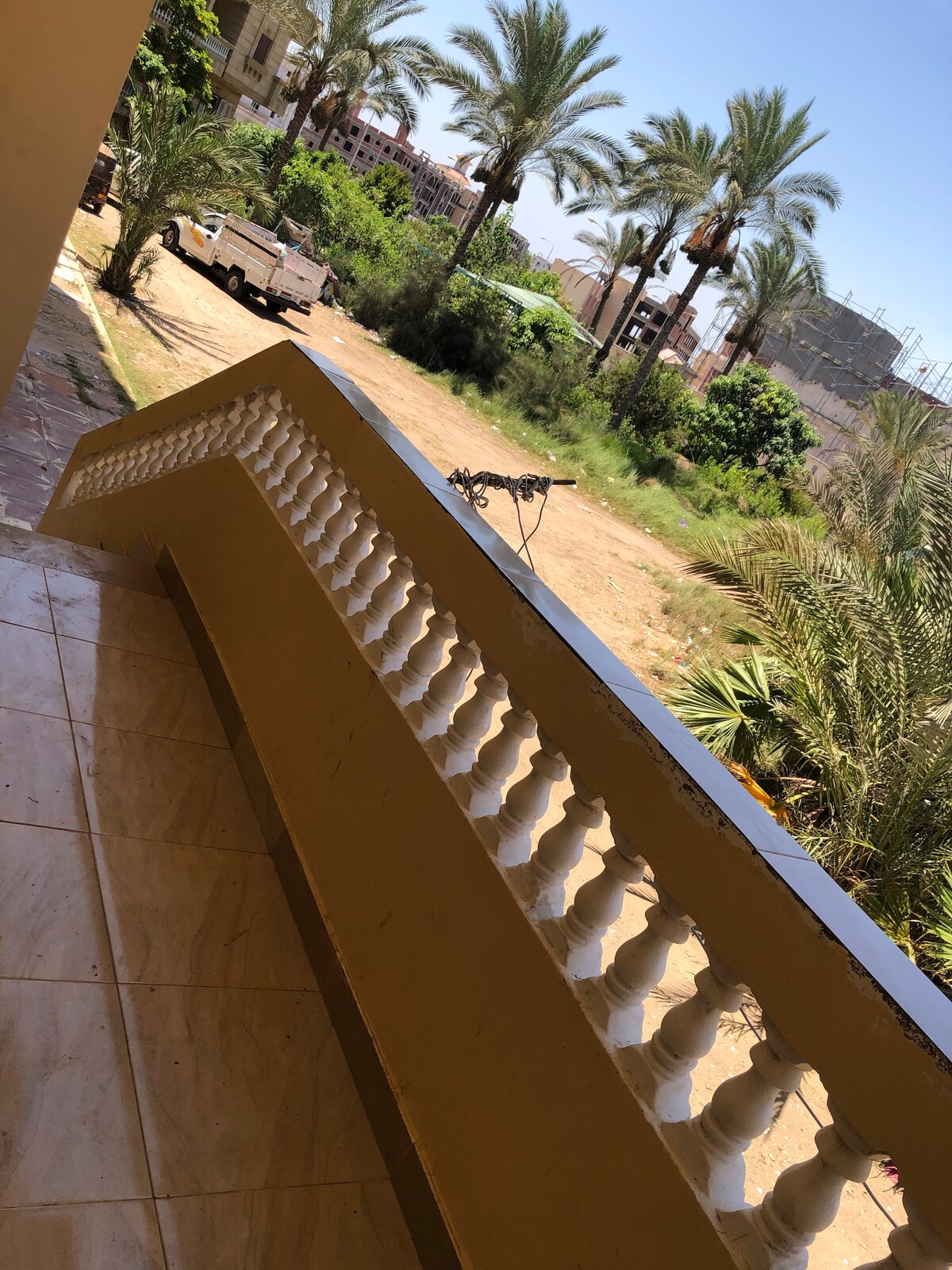 A view from a balcony showcases a sunlit outdoor area lined with palm trees. A staircase leads down to a garden space, with a vehicle parked on the gravel path. The railing, adorned with decorative balusters, frames the scene.