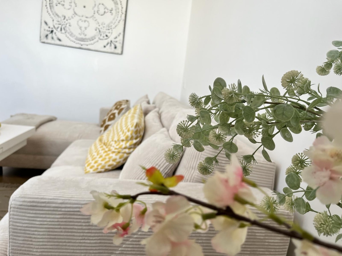 A modern living area is illuminated by natural light, featuring a plush sectional sofa adorned with decorative cushions. The scene includes a coffee table and a piece of wall art above the sofa, complemented by a delicate arrangement of faux flowers and greenery in the foreground.