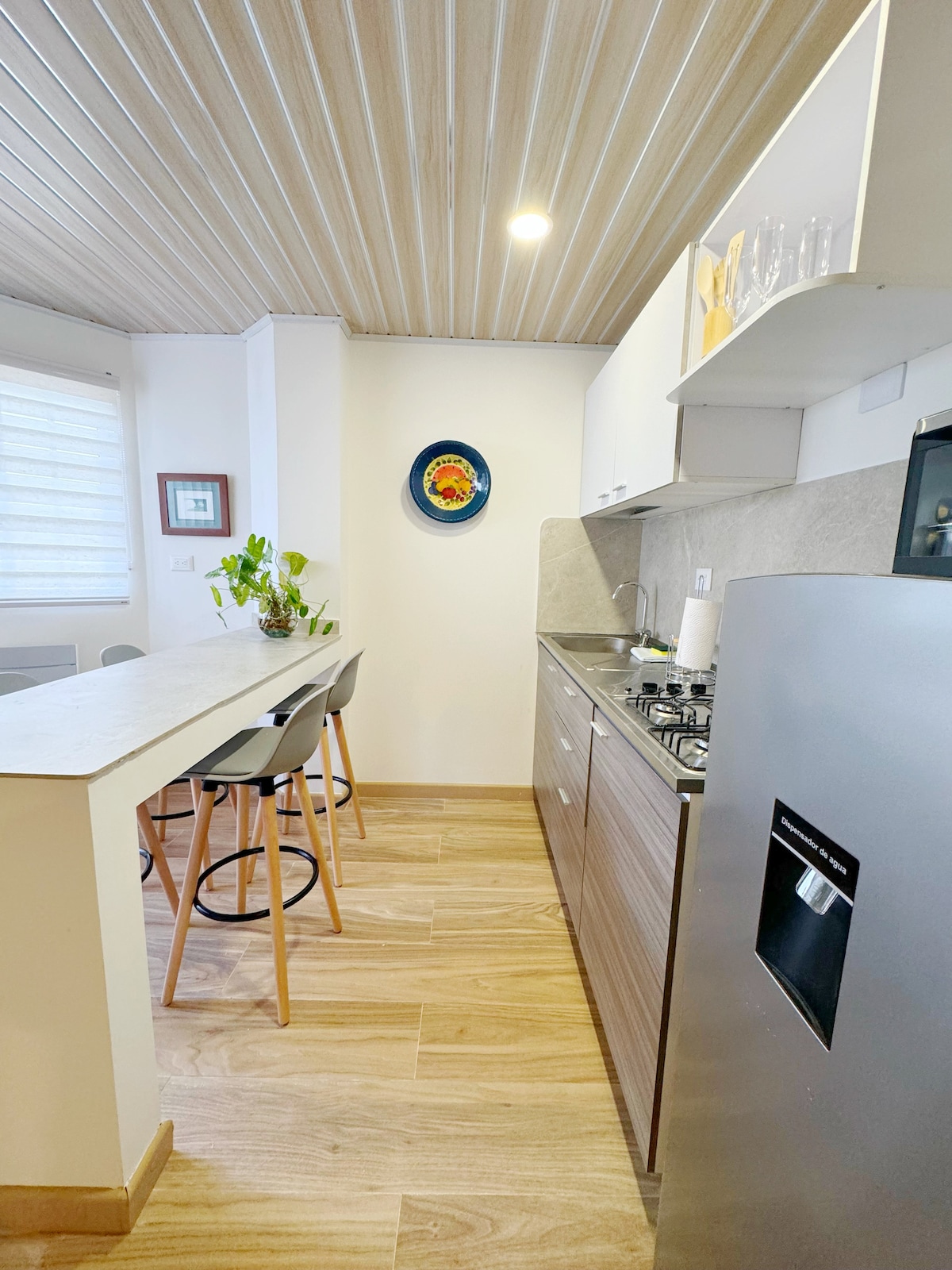 A modern kitchen is displayed, featuring a compact layout with sleek cabinetry and a stainless-steel refrigerator. A small dining area with three stools is positioned at a counter, complemented by a potted plant and decorative wall art, providing a functional and stylish space.
