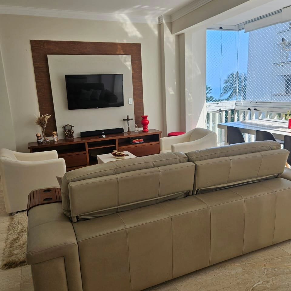 The living area features a comfortable sofa with side armrests, facing a sleek media console and flat-screen television. Natural light fills the space through large windows, while a blend of decorative items adds character to the room. A balcony with safety screens provides a view of the outside.