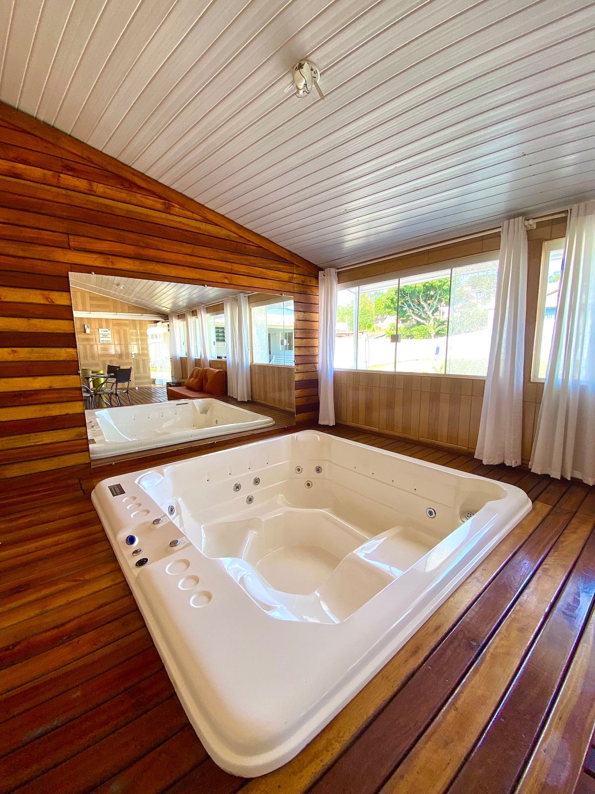 A spacious hot tub is positioned on wooden flooring, surrounded by a warm, natural wood-paneled wall. A large mirror reflects the hot tub and a bright window allows natural light to fill the area, enhancing the overall inviting ambiance.