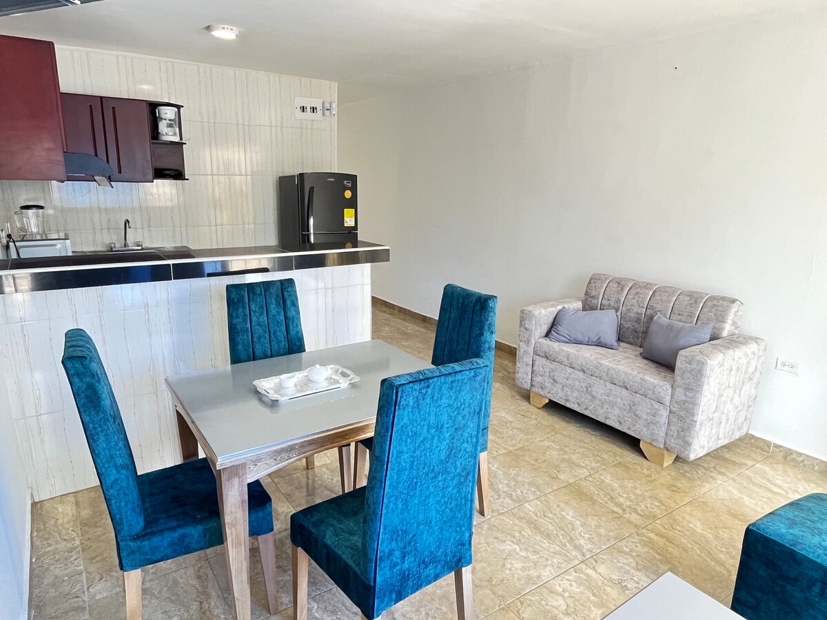 A modern kitchen area features sleek countertops and essential appliances, including a refrigerator. A dining table with four teal chairs is positioned next to a cozy gray sofa. The room is filled with natural light, enhancing the open and airy design.