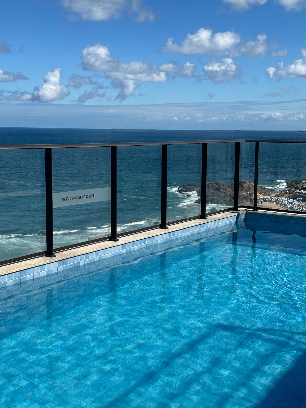 A rooftop pool is visible, bordered by glass railings that provide unobstructed views of the ocean. The clear water reflects the sky, which features scattered clouds and blue tones. A portion of the coastline can be seen in the distance.