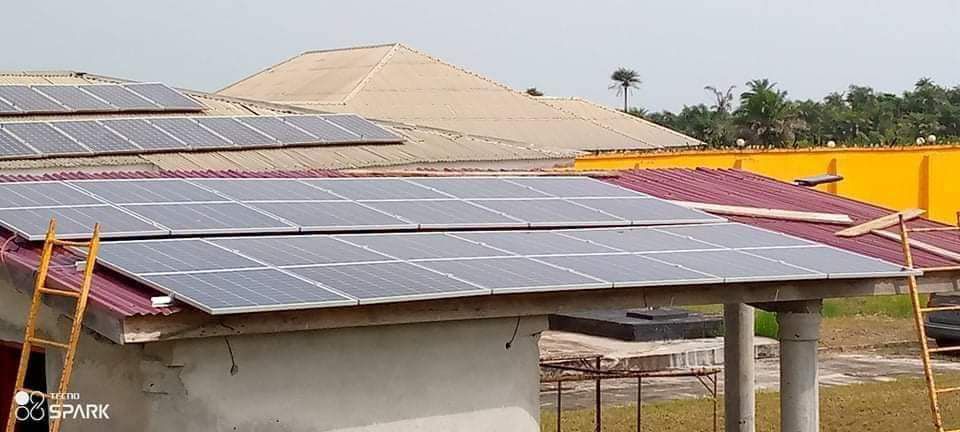 A rooftop is covered in rows of solar panels, reflecting light under a clear sky. The panels are mounted on a red pitched roof supported by concrete pillars, with construction materials visible in the background.