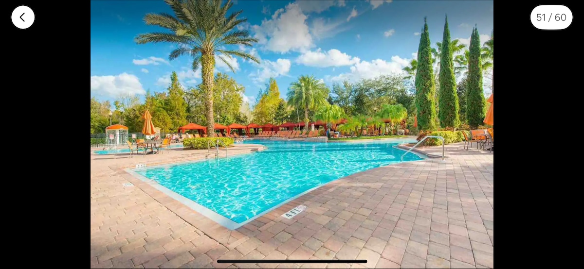 A large swimming pool is surrounded by palm trees and tropical landscaping. Lounge chairs are placed along the poolside, with orange umbrellas providing shade. The clear blue water reflects the sky, creating a serene atmosphere for relaxation.