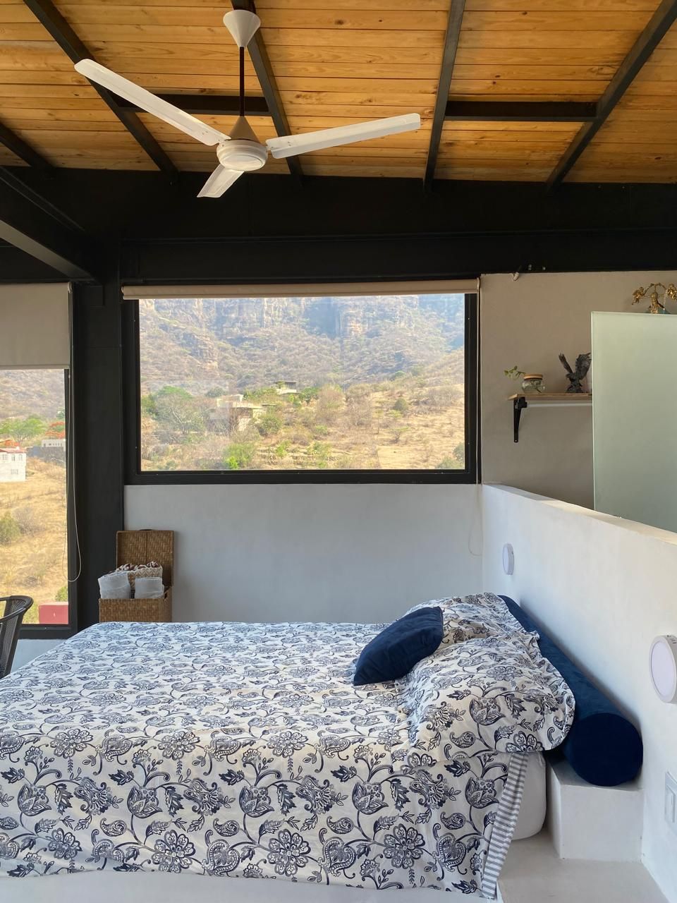 A serene bedroom is featured, showcasing a comfortable bed with a blue and white patterned blanket. Large windows provide a view of the surrounding mountain landscape. A ceiling fan and simple decor enhance the space, while minimal shelving adds functional character.