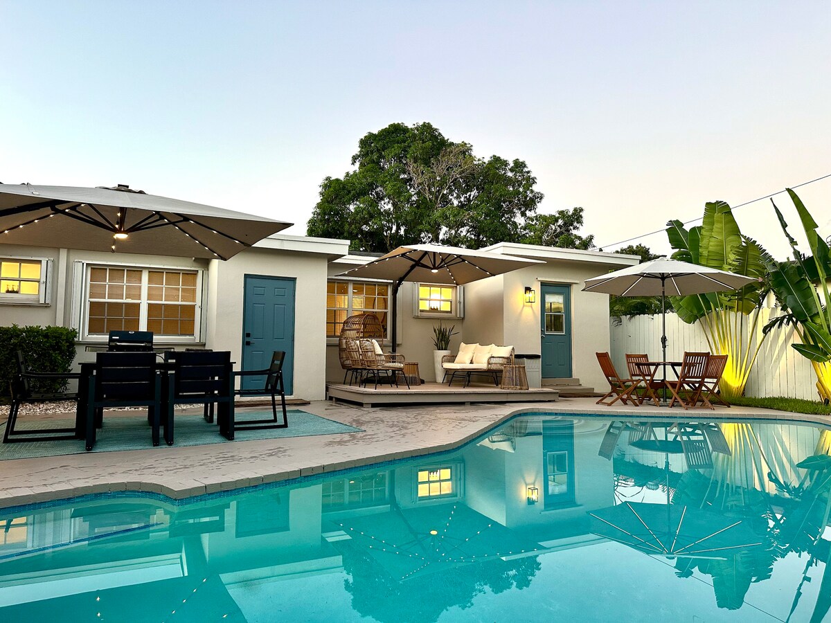 An inviting outdoor area features a pristine saltwater pool reflecting the surrounding greenery. Comfortable lounge chairs and a wooden dining set are shaded by large umbrellas. The backdrop presents a modern residence, with warm lighting highlighting the architecture and landscape.