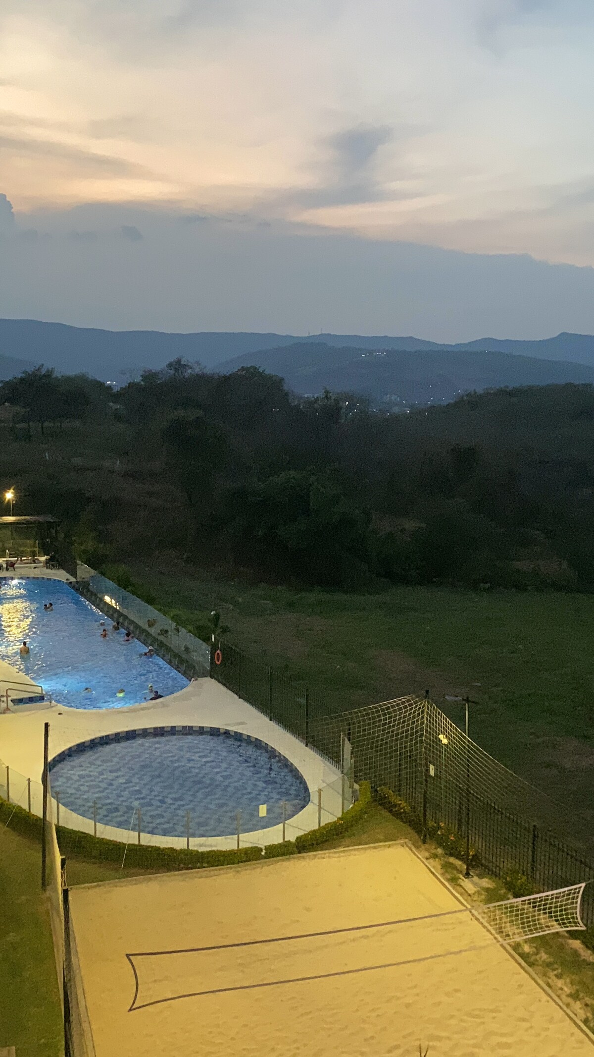 A serene outdoor pool area is highlighted, featuring a circular hot tub alongside a larger swimming pool. The backdrop includes rolling green hills under a softly lit sky. A sandy area appears in the vicinity, with a volleyball net present.