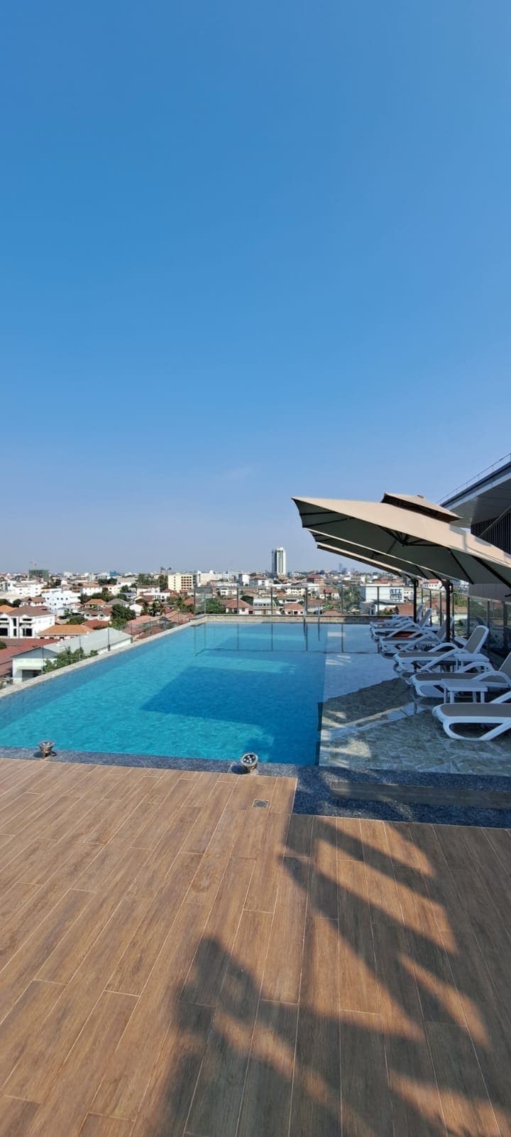 An infinity pool extends towards a panoramic city view under a clear blue sky. Comfortable lounge chairs are lined along the poolside, while shaded areas provide respite from the sun. The terrace features wooden decking, complementing the tranquil waters of the pool.