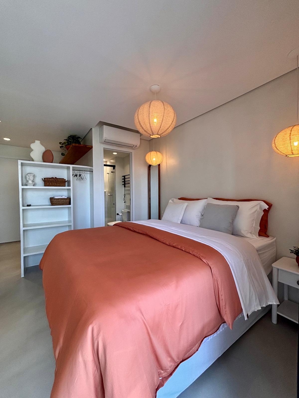 A modern sleeping area features a spacious bed with soft, coral-colored bedding and decorative cushions. Warm ambient lighting is provided by pendant lamps, while built-in shelves display tasteful accents. A pathway leads toward a stylish bathroom and additional living space.