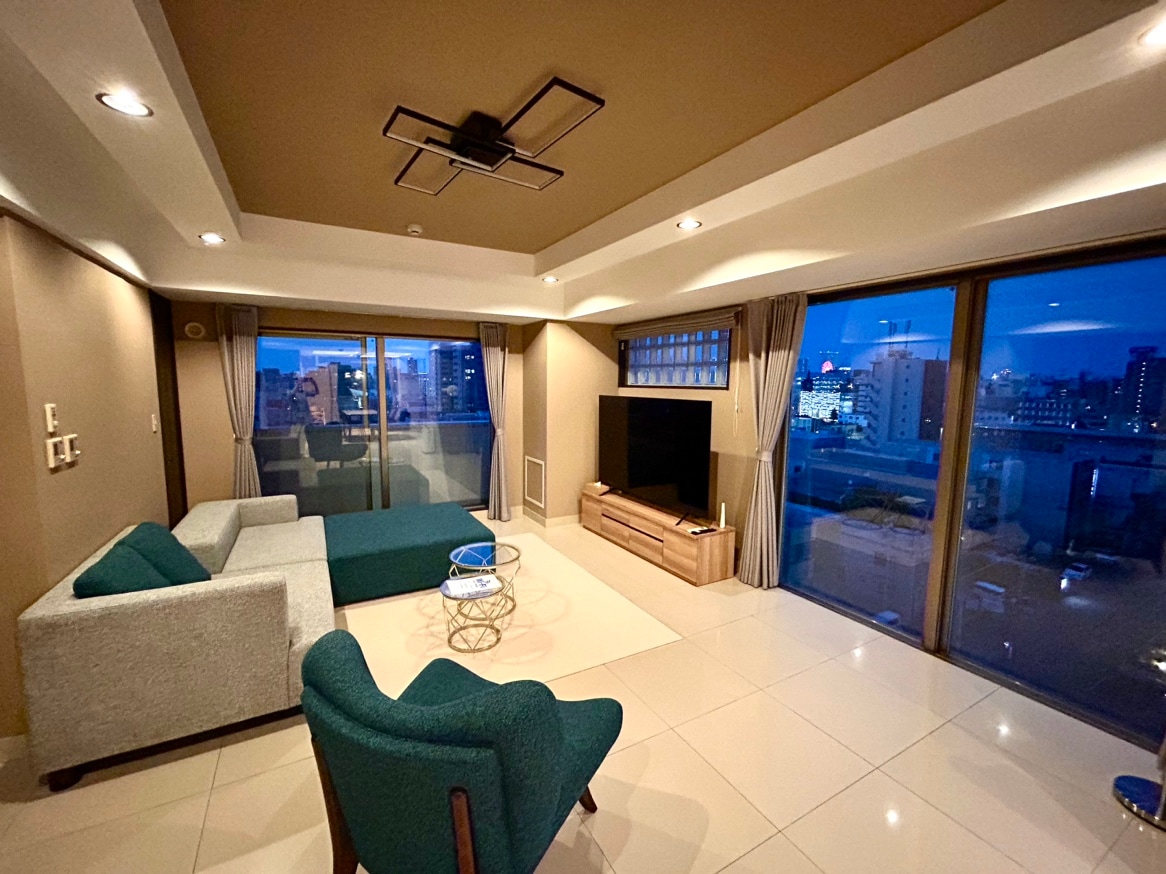A spacious living area features large windows offering views of the city skyline. A light-colored sofa and a deep blue accent chair are arranged around a modern coffee table. Soft lighting highlights the neutral tones and clean lines of the design.