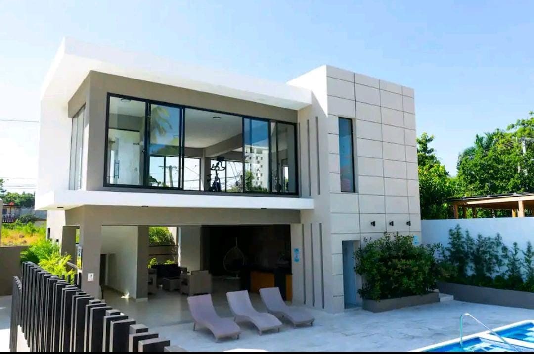 A modern two-story home is showcased with large glass windows, allowing natural light to fill the interior. A patio extends from the structure, featuring multiple lounge chairs beside a pool. Lush greenery surrounds the property, enhancing the outdoor space.