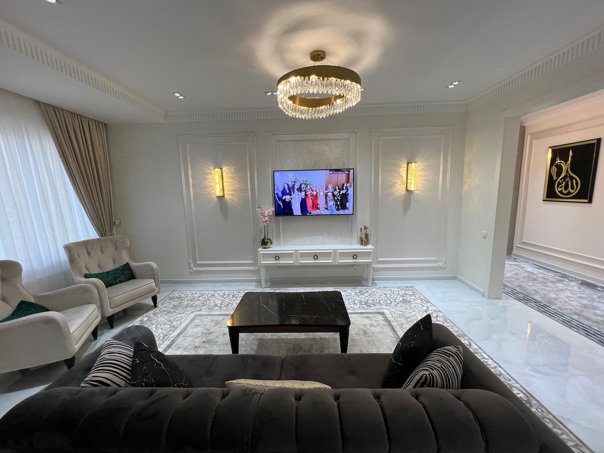 A living area is presented with a plush dark sofa adorned with striped cushions. A black coffee table is centered on a light area rug. Two elegant armchairs flank the sofa, while a wall-mounted television and decorative wall sconces further enhance the space's ambiance.