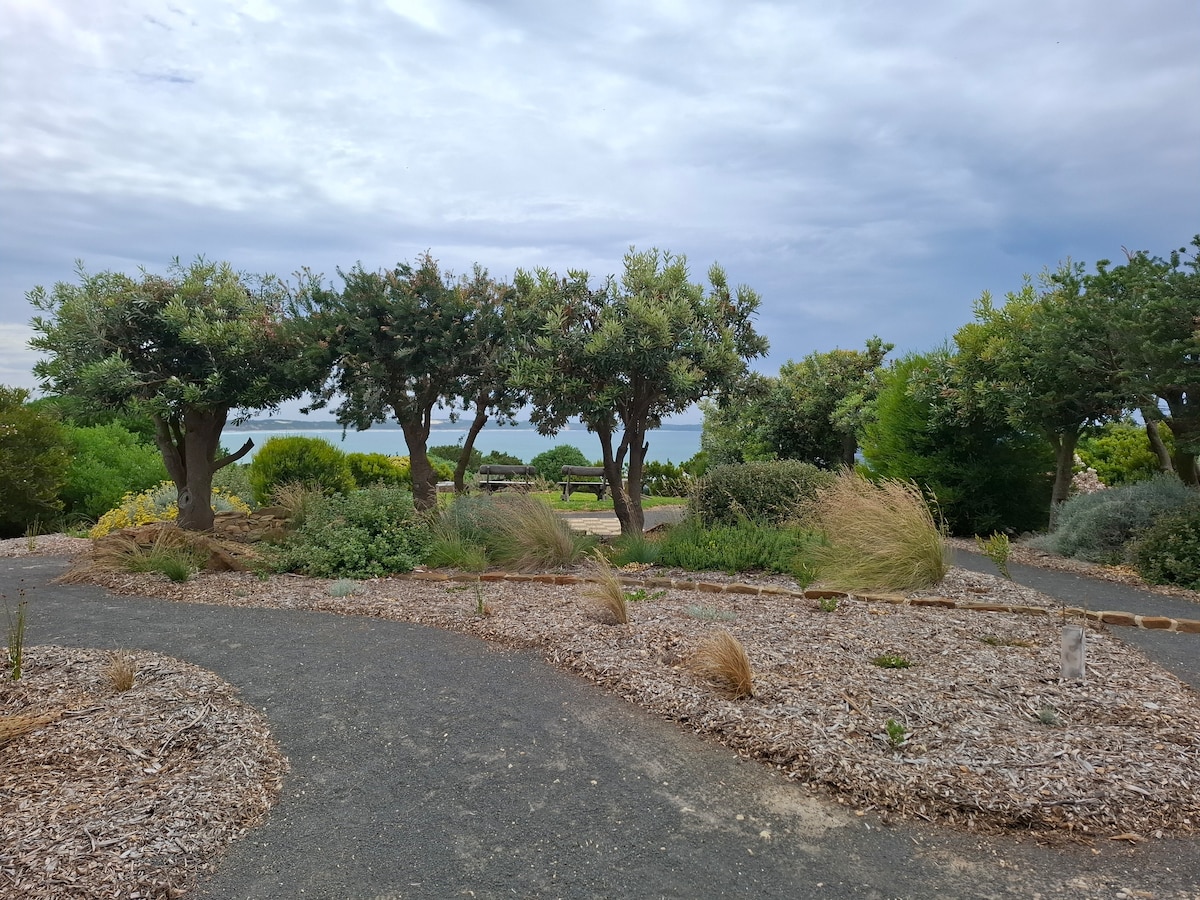 A landscape featuring winding paths bordered by low shrubs and clusters of trees is presented. Natural grasses sway gently in the breeze, while patches of greenery create a serene outdoor space. The scene suggests a tranquil setting with fields in the backdrop.