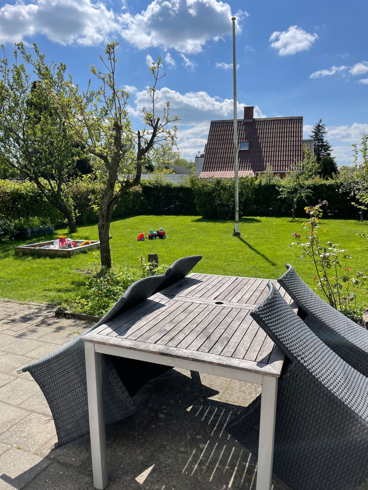 An outdoor area is presented with green grass and a wooden table surrounded by four woven chairs. A sandbox is visible in the distance, alongside scattered toys that add to the inviting outdoor environment. Sunlight enhances the overall appeal of the space.