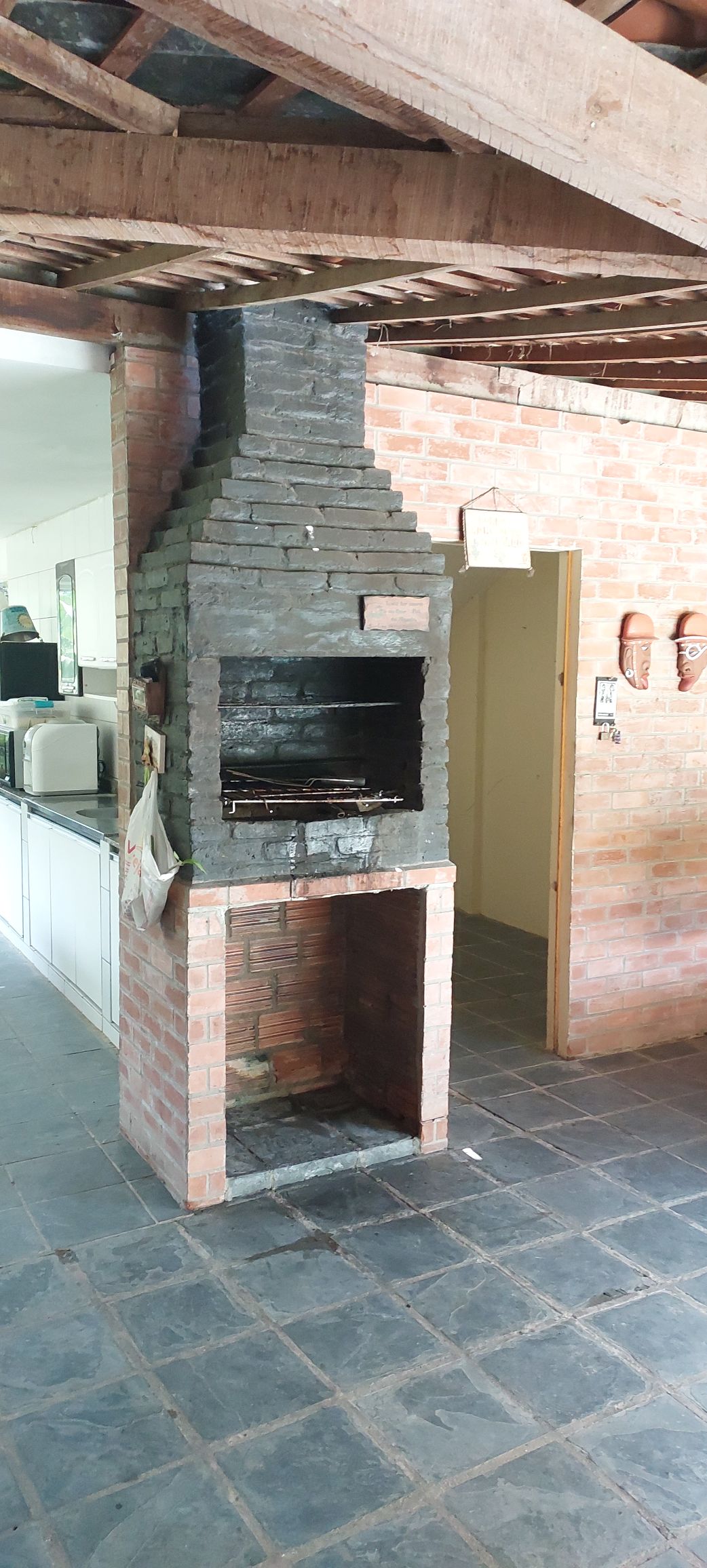 A brick barbecue grill is positioned in a rustic kitchen area, featuring a blackened chimney and an open grill space beneath. The surrounding walls are adorned with warm, earthy tones, and a spacious countertop is visible to one side, adding functionality to the inviting cooking space.