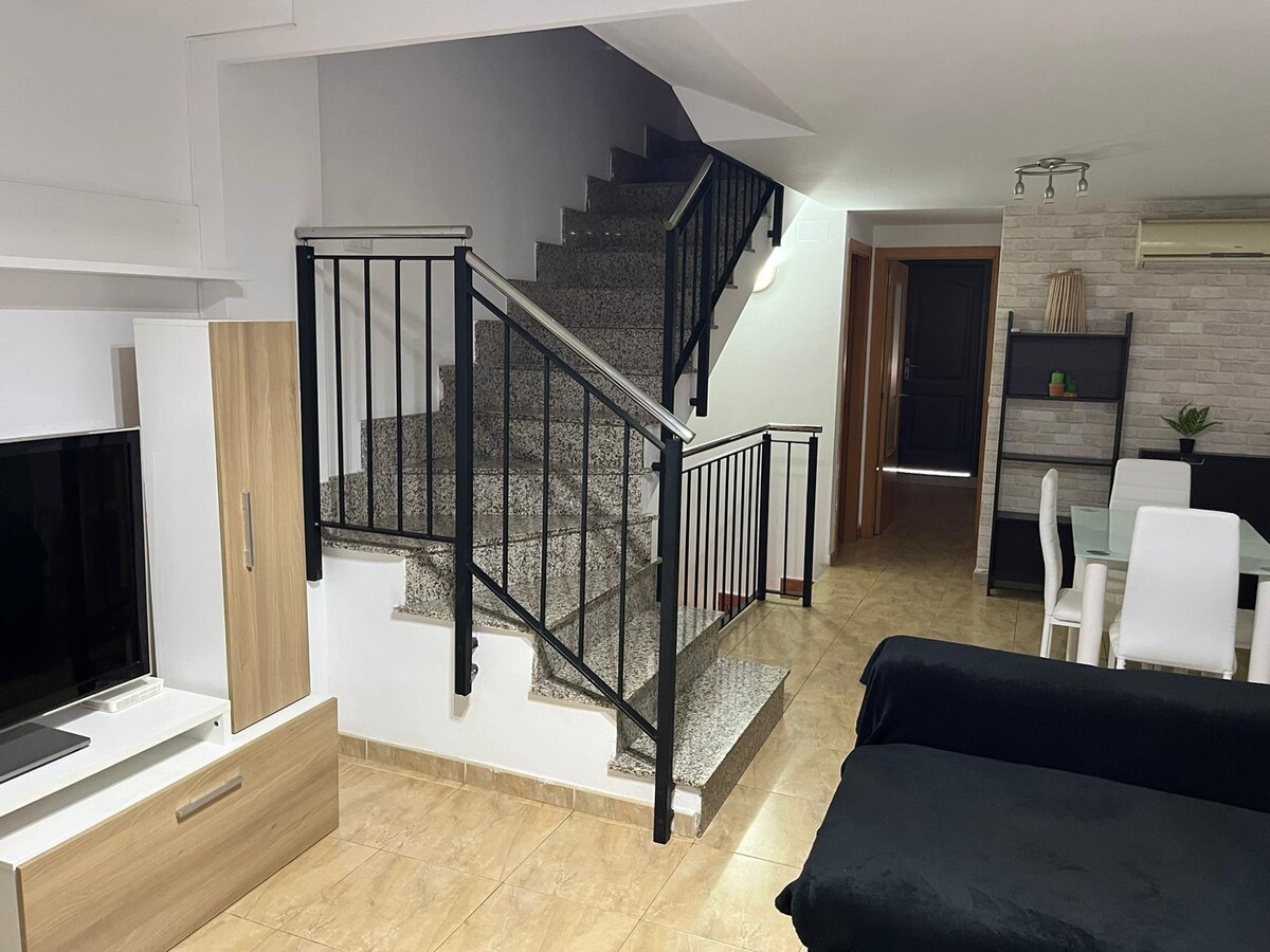 A well-lit living area is visible, featuring a staircase leading to the upper level. The space includes a television mounted on a light wood unit and a modern dining area with white chairs. The floor is tiled in a warm beige tone.
