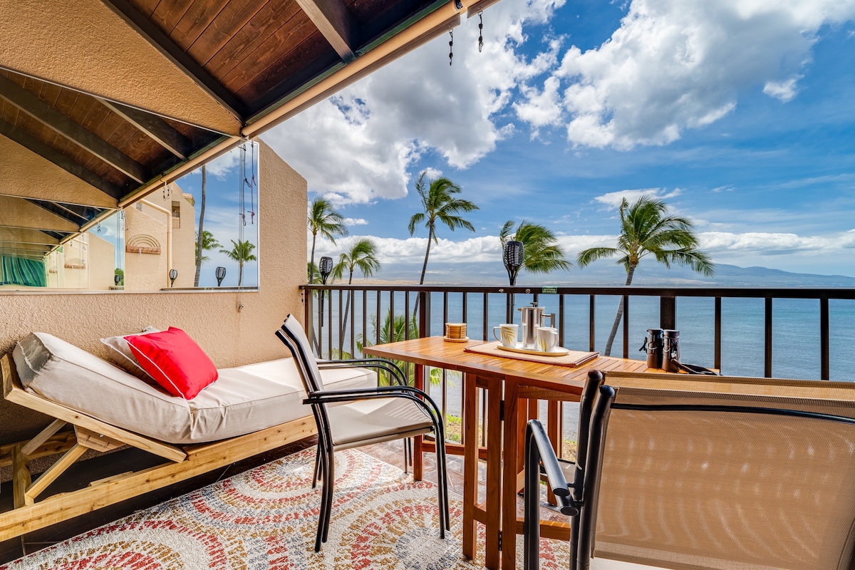 A private balcony is depicted, featuring a wooden table and two chairs alongside a lounge chair with a red cushion. The ocean and palm trees are visible in the background under a partly cloudy sky, creating a calming coastal scene.