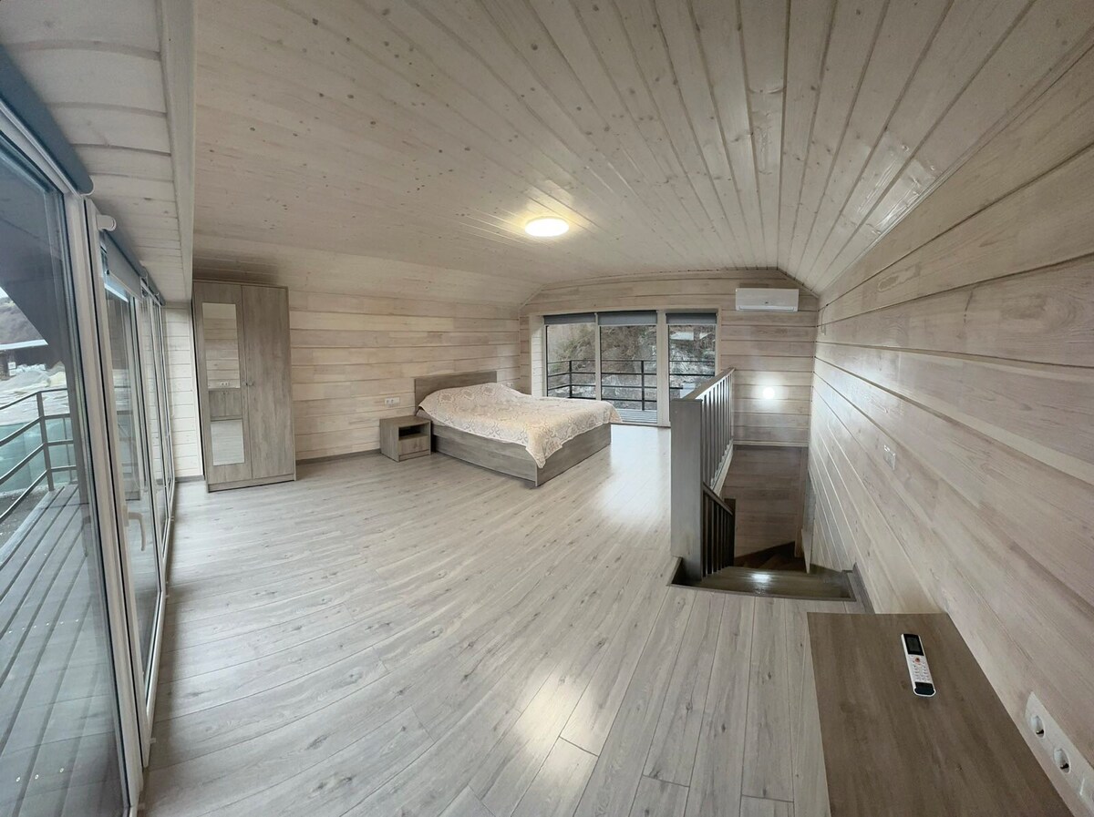 An airy sleeping area features wooden paneling and a vaulted ceiling. A spacious bed is positioned near large windows, allowing natural light to fill the room. A wooden dresser and a small nightstand accompany the bed, while stairs lead to the lower level.