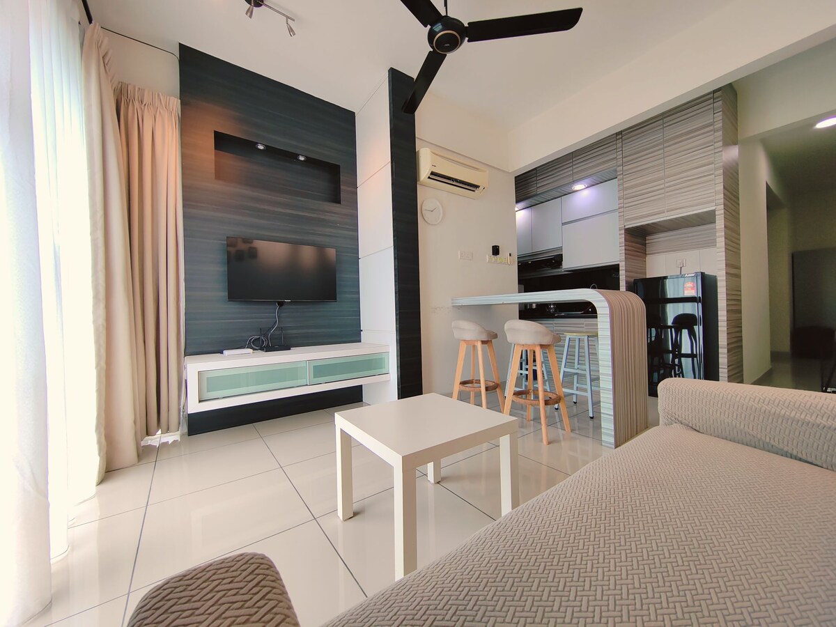 A modern living area features a flat-screen television mounted on a textured wall. A light-colored sofa faces a sleek coffee table, with natural light filtering through sheer curtains. The open concept connects seamlessly to a dining area with barstools and a kitchen in the background.
