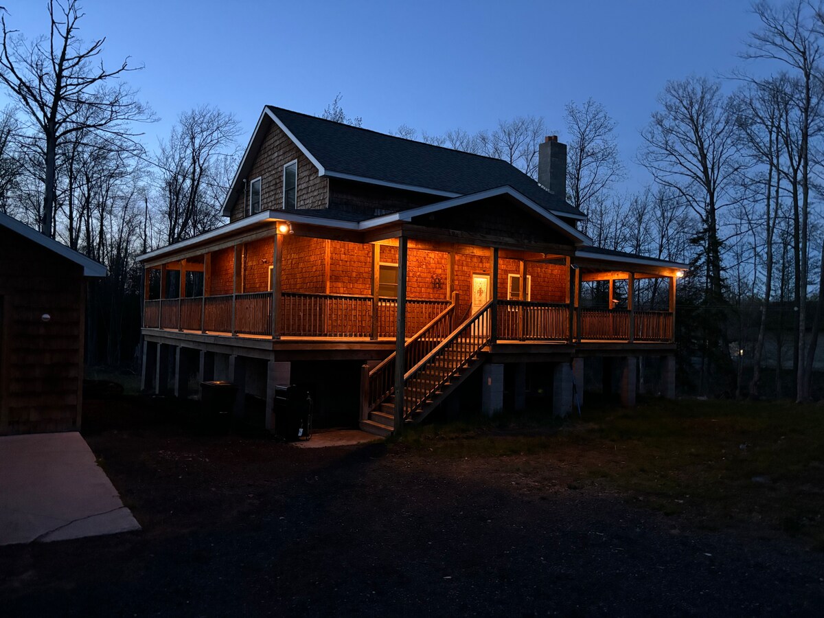 The exterior of the house is illuminated in warm light, showcasing a large wrap-around porch with steps leading up to the entrance. Surrounding trees provide natural textures, and the setting is highlighted by the early evening sky.