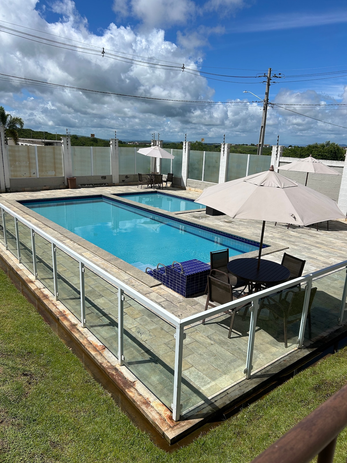 A spacious outdoor area features a clear swimming pool, surrounded by a protective glass railing. Two umbrella-covered tables with chairs and a lounge area are visible, offering a spot for relaxation. Lush greenery is present in the surrounding landscape, under a bright blue sky with scattered clouds.