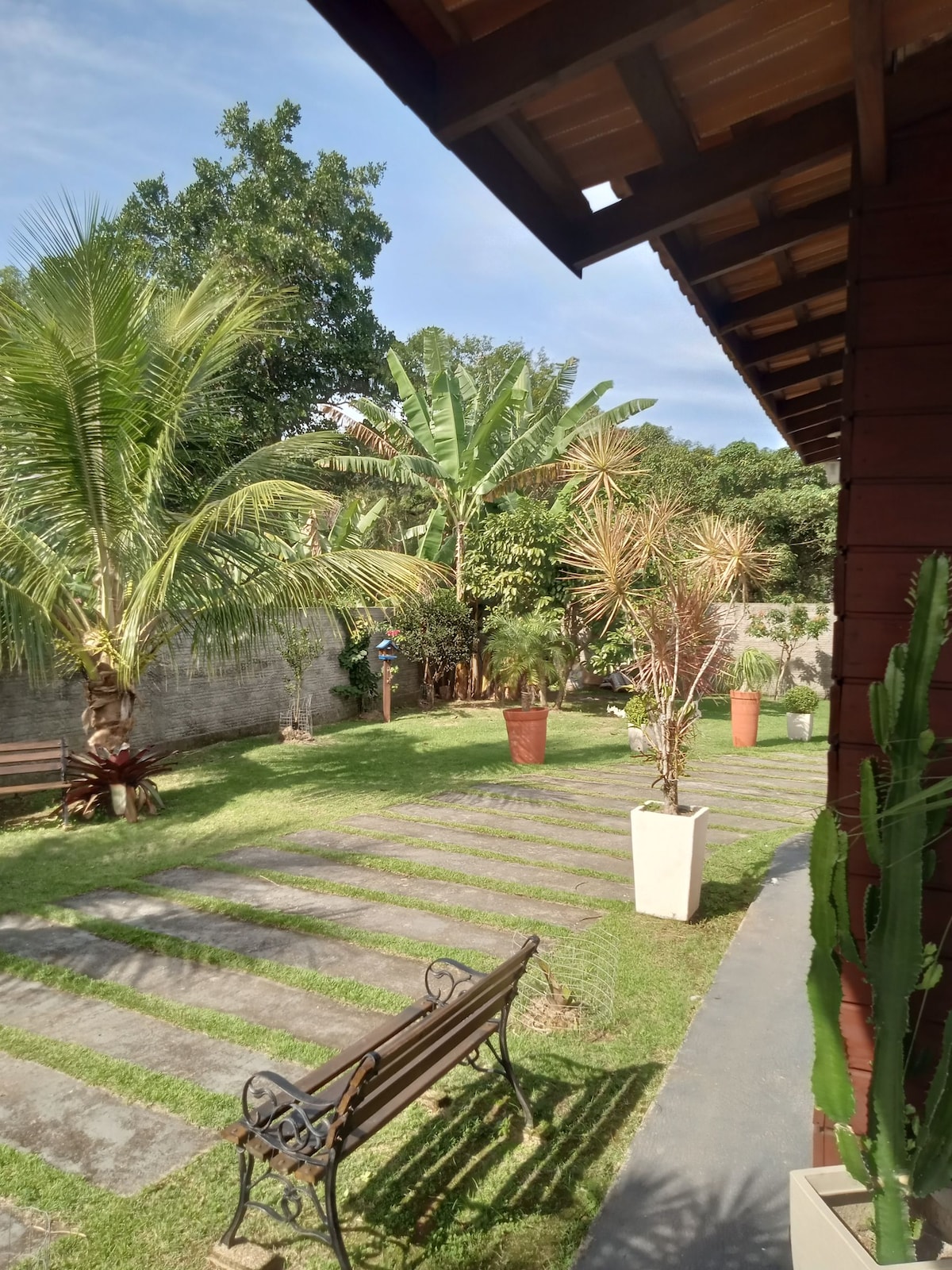 A well-maintained garden is presented with a combination of tropical plants and trees, featuring stone pathway strips leading through the lush green grass. Benches are positioned along the path, inviting relaxation amidst the tranquil surroundings.