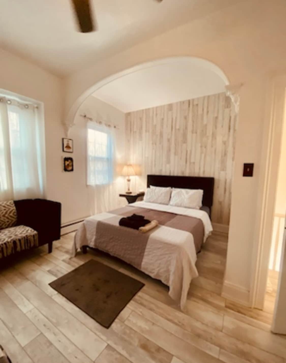 A inviting room features a spacious bed with soft linens and a cozy blanket. Natural light filters through two large windows, illuminating the neutral walls with wooden accents. A comfortable seating area is also visible, adding to the room's welcoming environment.