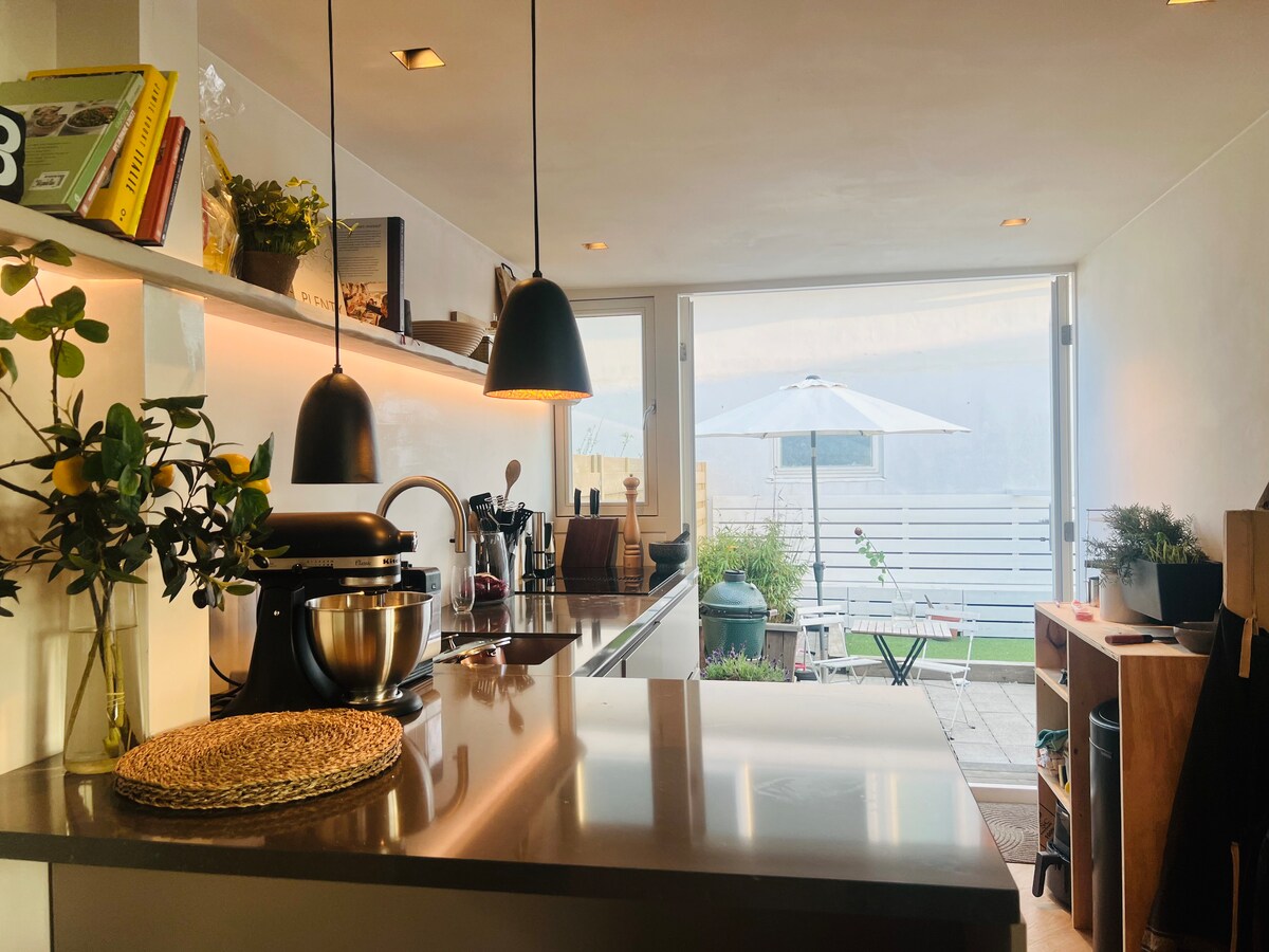 A modern kitchen space is showcased with sleek countertops and overhead pendant lighting. Cooking utensils and appliances are neatly arranged, while a view of the outdoor patio with an umbrella and greenery is visible through large glass doors, creating a light-filled atmosphere.